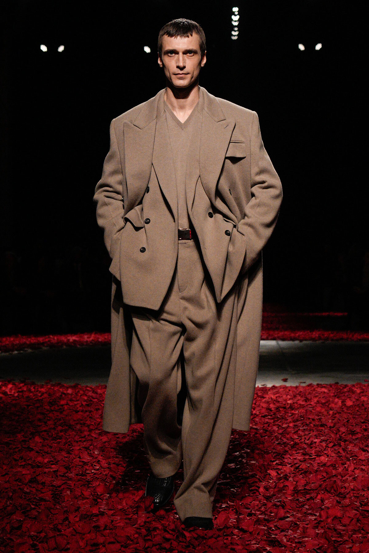 Male model walking a fashion runway in a tan suit with a long overcoat, on a red-petaled catwalk backdrop.