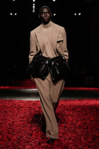 Male model on a fashion runway wearing a tan suit and turtleneck, paired with oversized black belt bags.