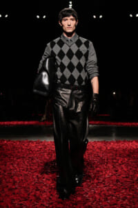 Male model on a runway wearing a gray argyle sweater, black leather pants and gloves, carrying a large black leather bag.