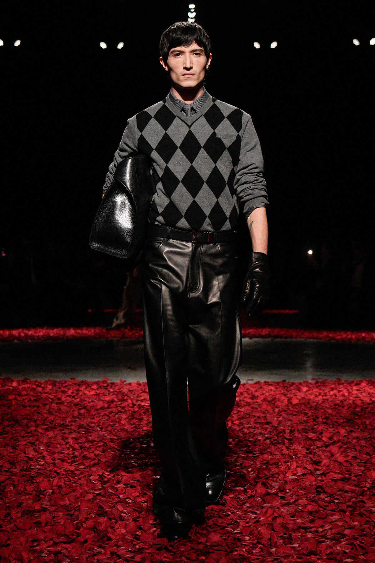 Male model on a runway wearing a gray argyle sweater, black leather pants and gloves, carrying a large black leather bag.