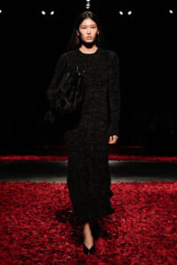 Model walks a runway in a black textured knit dress, carrying a large black bag; red petals cover the floor beneath her.