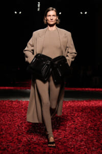 Runway model in a taupe coat and matching ensemble, holding two large black leather handbags at the front amid a red-petaled runway.
