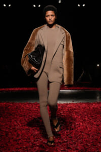 Model walking a runway in a neutral-toned suit with a brown fur shawl, holding a black bag, on a red petal-covered floor.