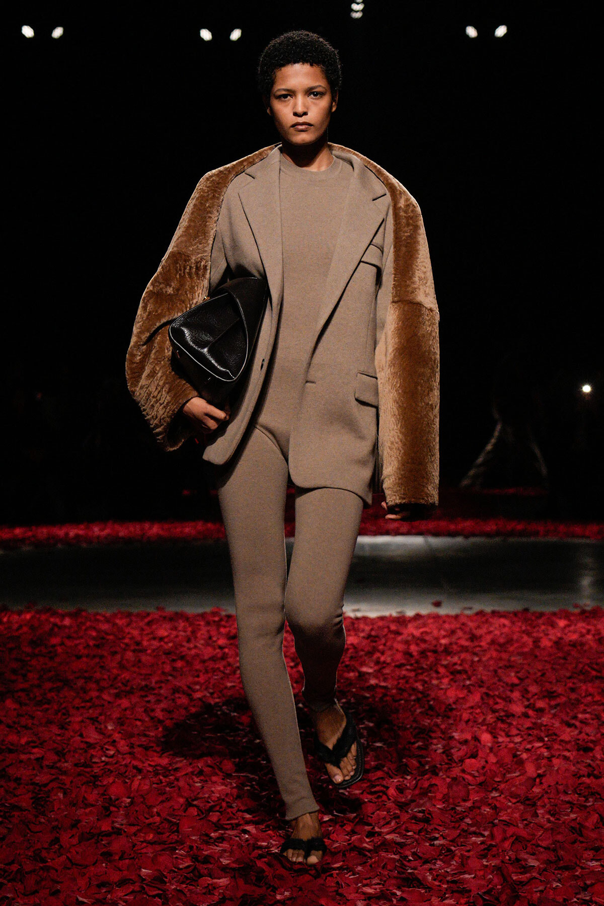 Model walking a runway in a neutral-toned suit with a brown fur shawl, holding a black bag, on a red petal-covered floor.