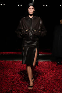 Model walking a runway in a dark brown leather bomber jacket and matching slit skirt amid red petals on the floor