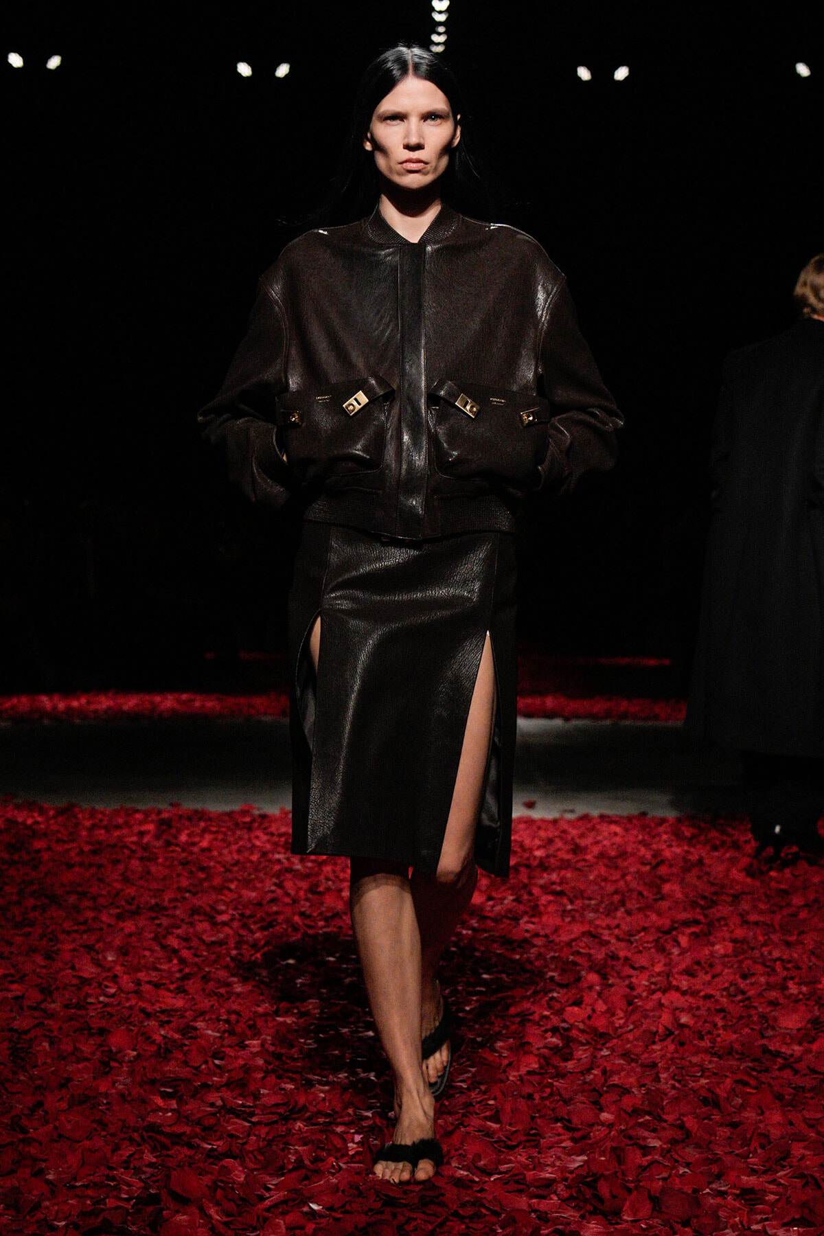 Model walking a runway in a dark brown leather bomber jacket and matching slit skirt amid red petals on the floor