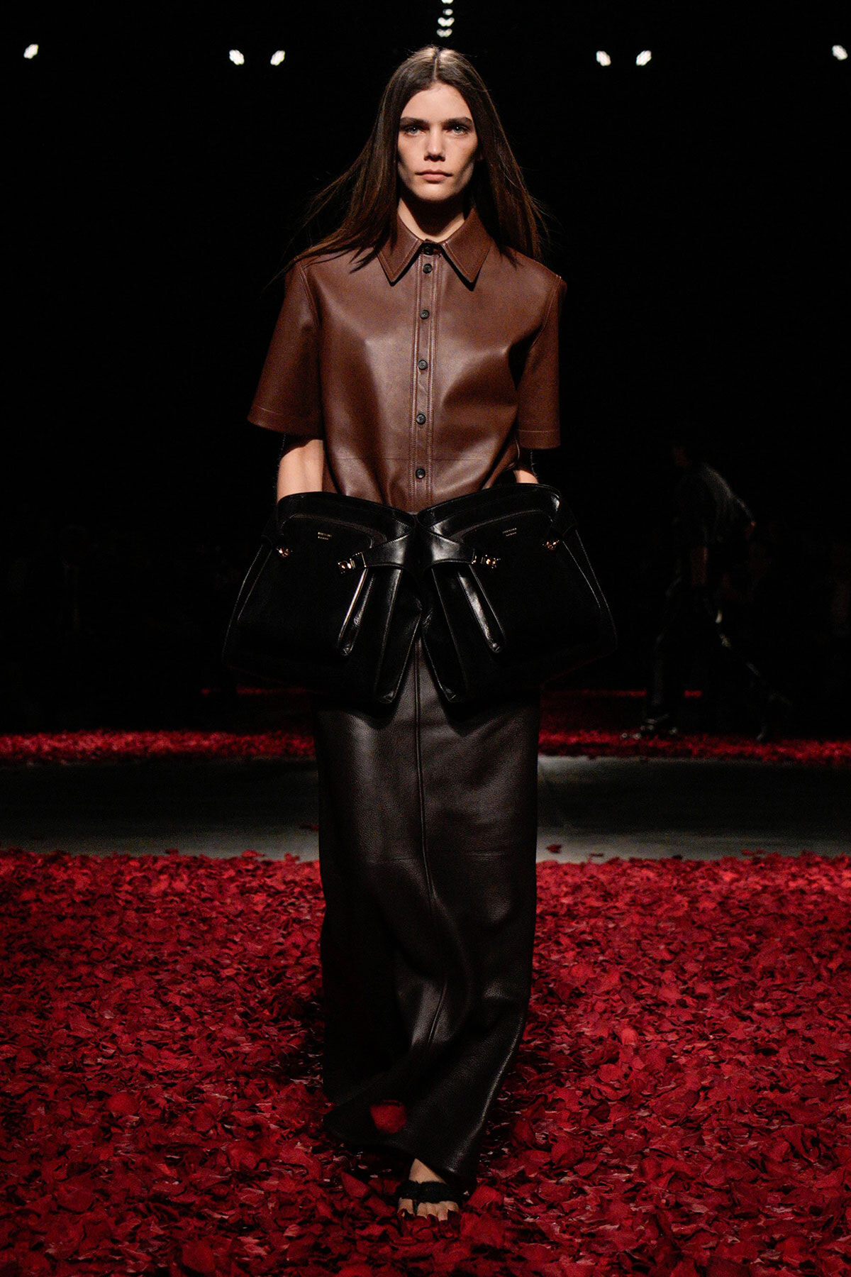 Model on a runway wearing a brown leather short-sleeve shirt and black leather wide-leg pants, carrying a large black handbag with buckles.