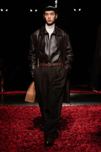 Male model on a runway wearing a dark brown leather jacket over a white turtleneck, with black trousers and a tan bag amidst red petals.