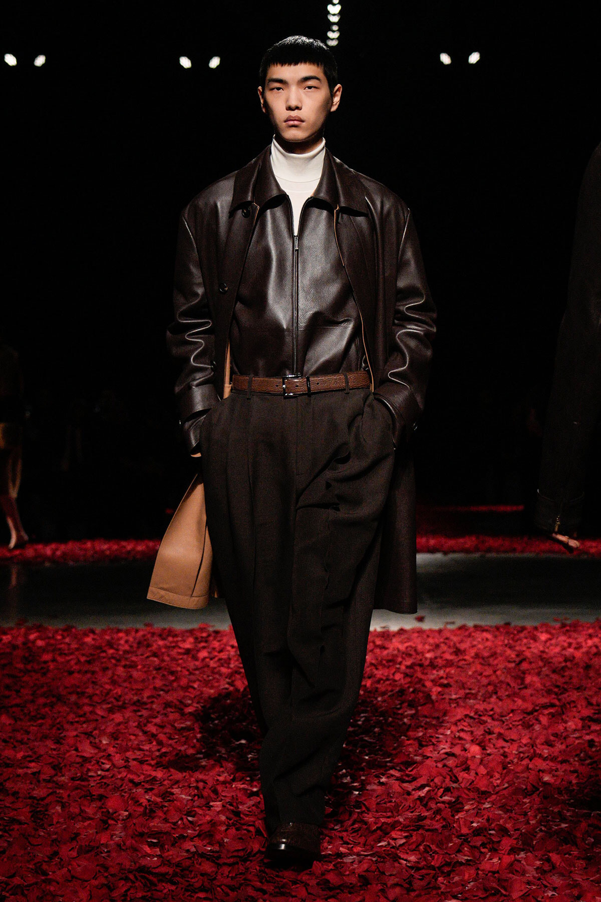 Male model on a runway wearing a dark brown leather jacket over a white turtleneck, with black trousers and a tan bag amidst red petals.