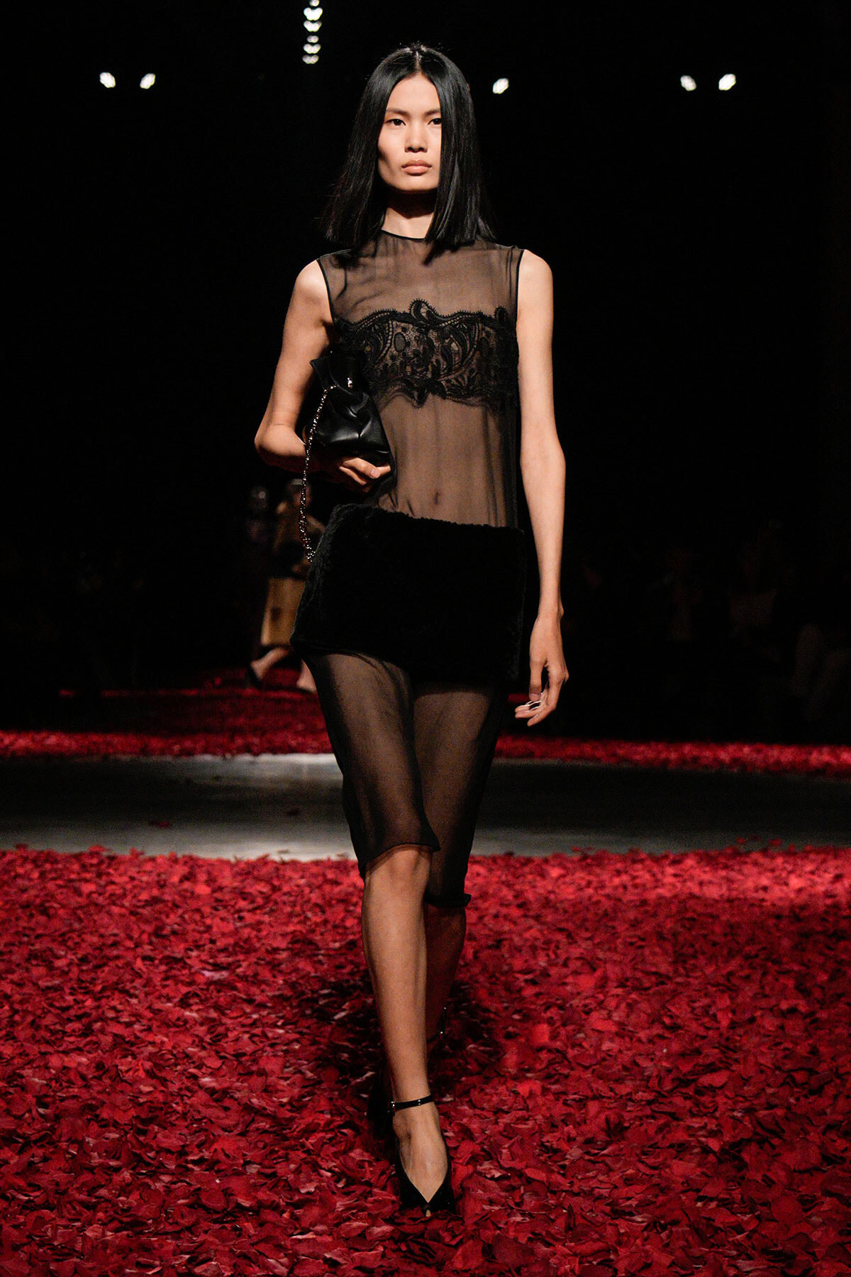 Model walks a runway in a sheer black lace dress, carrying a small black clutch, on a red petal-covered floor.