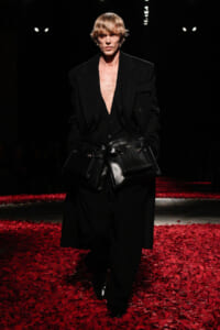 Male model on a runway wearing a black oversized blazer and matching pants, with multiple black leather pouches strapped around the waist. Red petals on the floor.”] ,