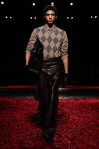 Male fashion model on a dark runway wearing a beige-gray argyle sweater, brown leather trousers, black gloves, and carrying a large black leather bag.