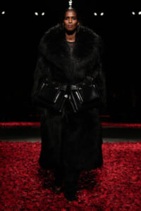 Runway model in a black fur coat with a large belt of black bags, walking on a red-petal floor at a fashion show