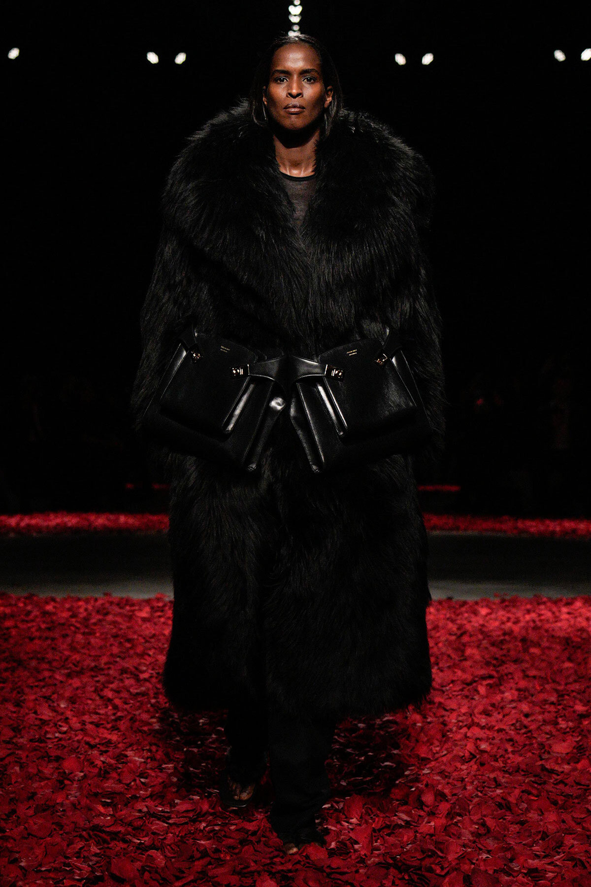 Runway model in a black fur coat with a large belt of black bags, walking on a red-petal floor at a fashion show