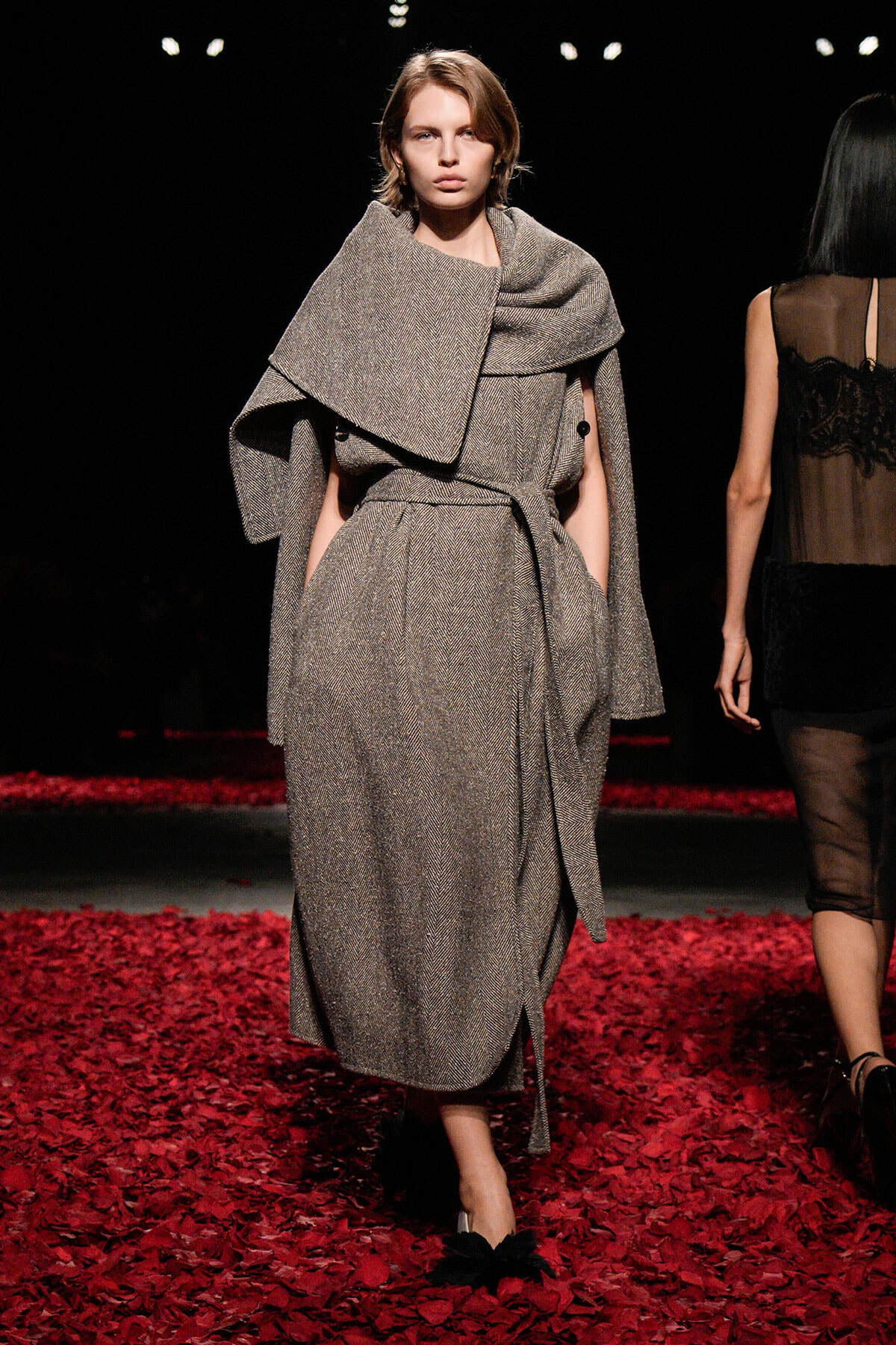 Model walking a runway in a gray textured wrap coat with a large draped collar, belted at the waist, on a red-petaled catwalk.