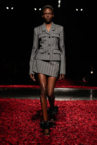Model walking a fashion runway in a gray pinstripe tailored blazer and mini skirt among red rose petals.