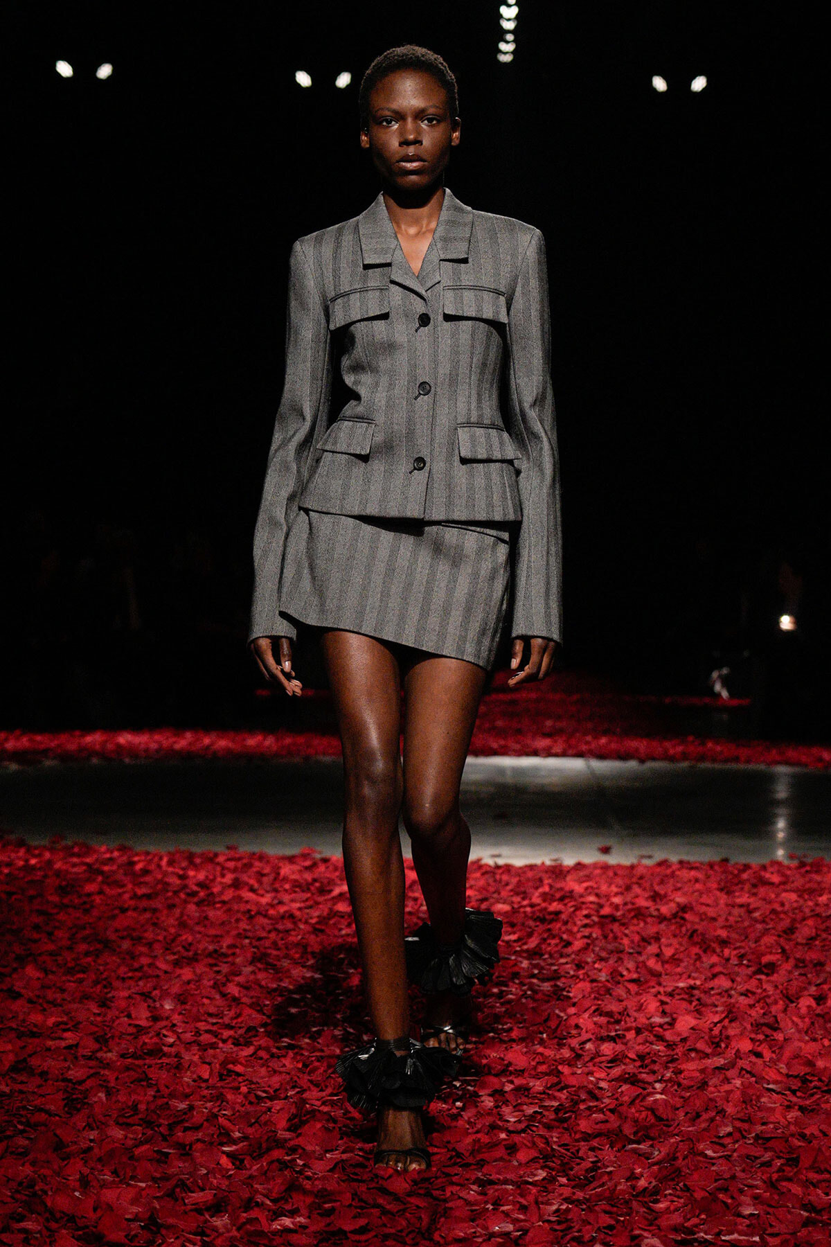 Model walking a fashion runway in a gray pinstripe tailored blazer and mini skirt among red rose petals.