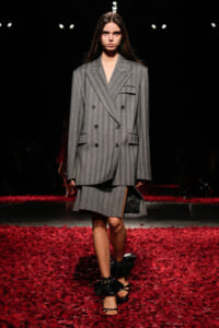 Model walks a fashion runway in an oversized gray pinstripe double-breasted suit with a matching skirt, black ruffled sandals, red petals on the floor.