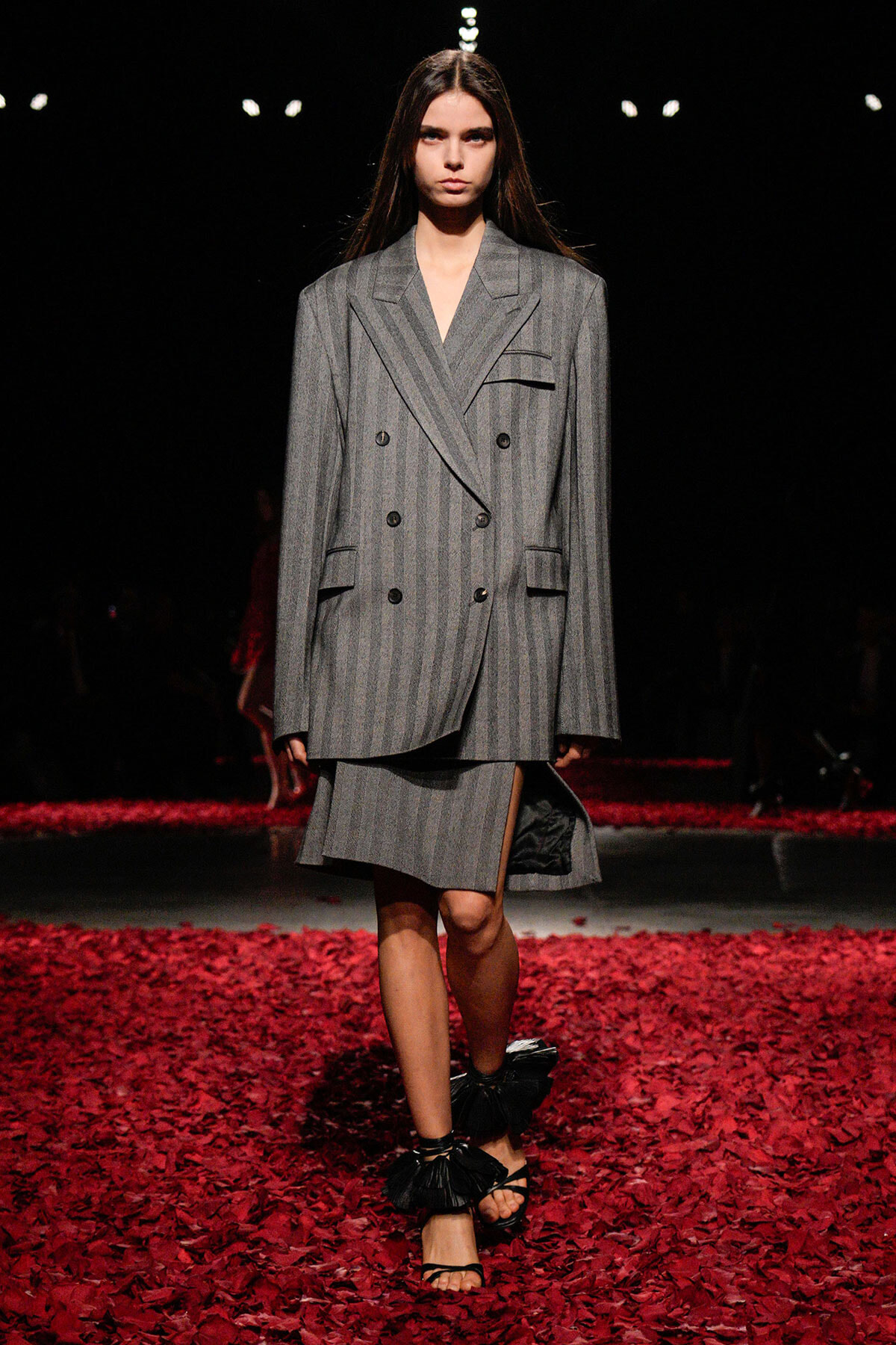 Model walks a fashion runway in an oversized gray pinstripe double-breasted suit with a matching skirt, black ruffled sandals, red petals on the floor.