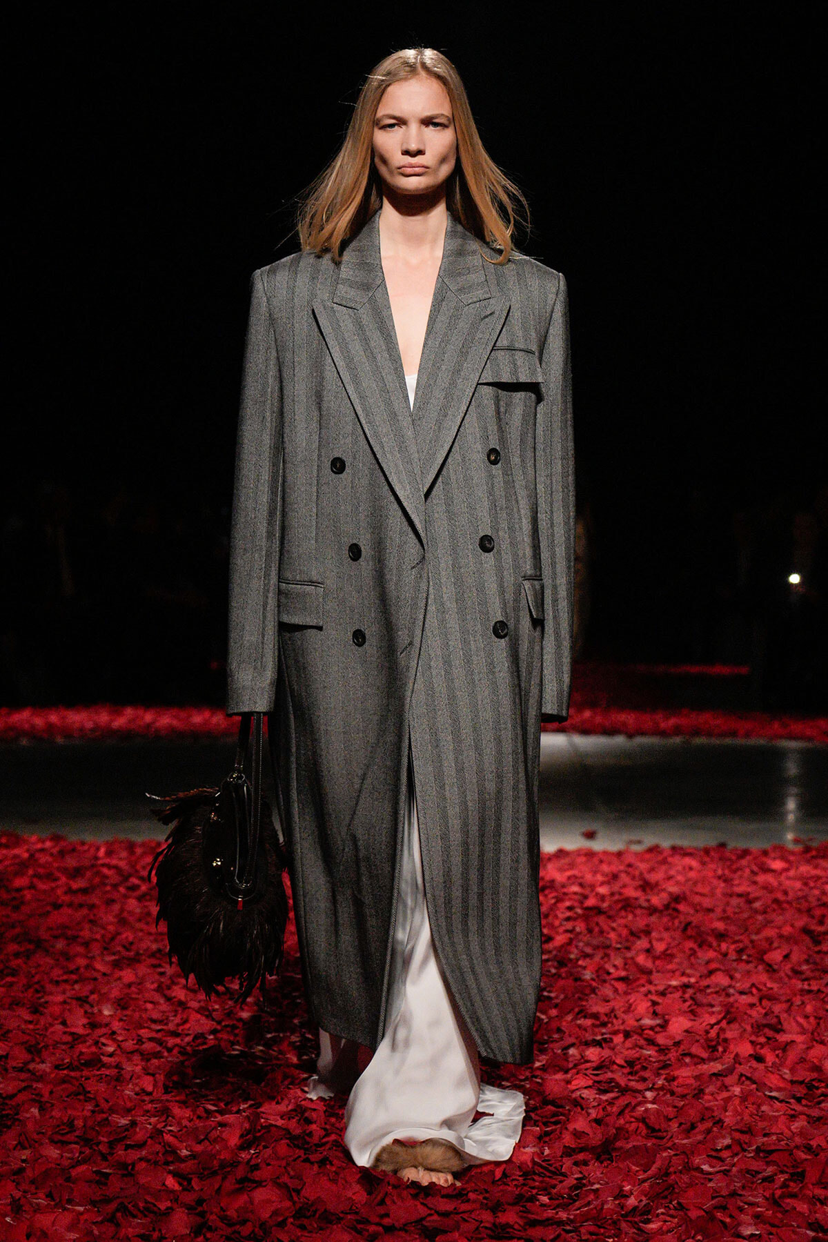 Model on a runway wearing a long gray pinstripe double-breasted coat, white dress, and black feathered handbag, walking over red petals.