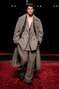 Male model walks a fashion runway wearing an oversized gray herringbone suit with a long coat, on a red-petaled runway.
