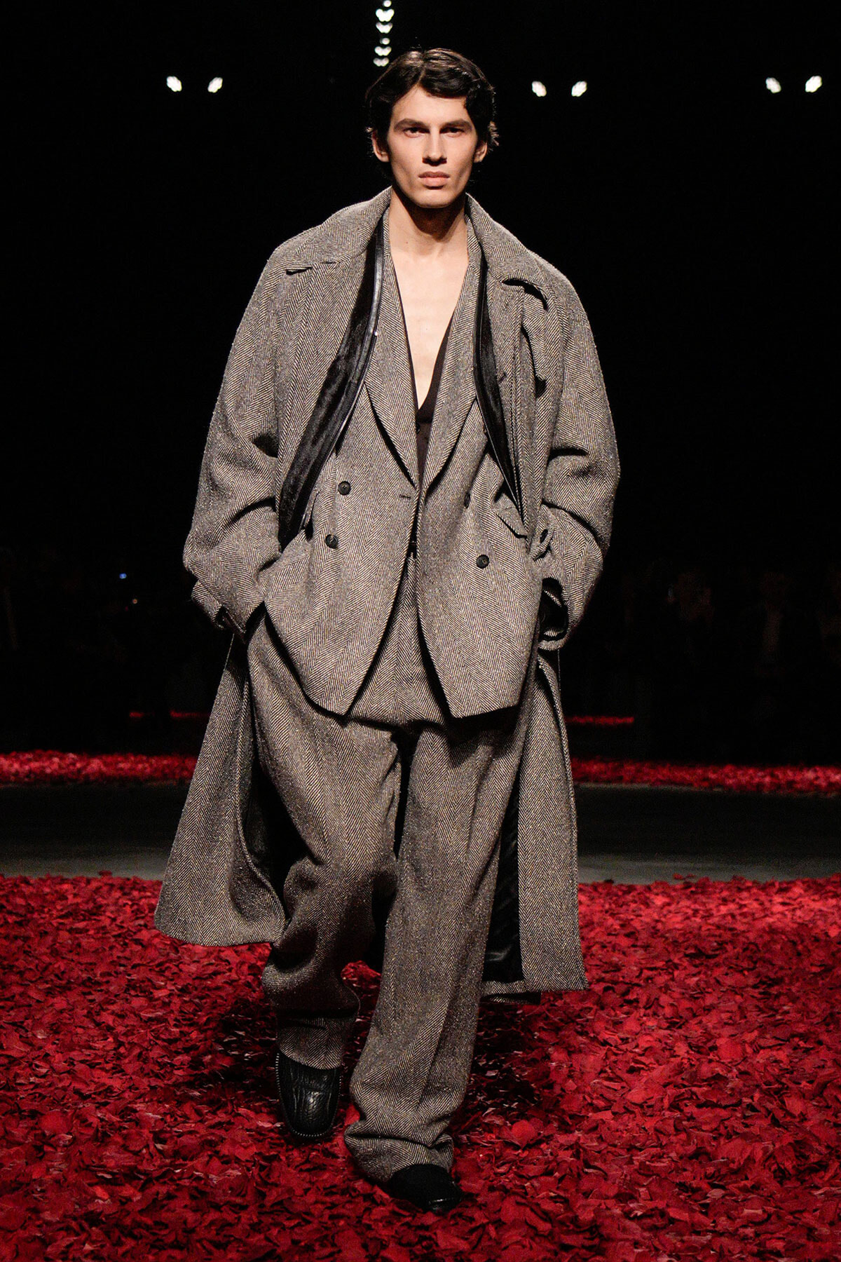 Male model walks a fashion runway wearing an oversized gray herringbone suit with a long coat, on a red-petaled runway.
