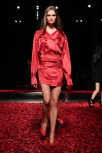 Model walking the runway in a red satin trench dress with belt on a petal-strewn floor.