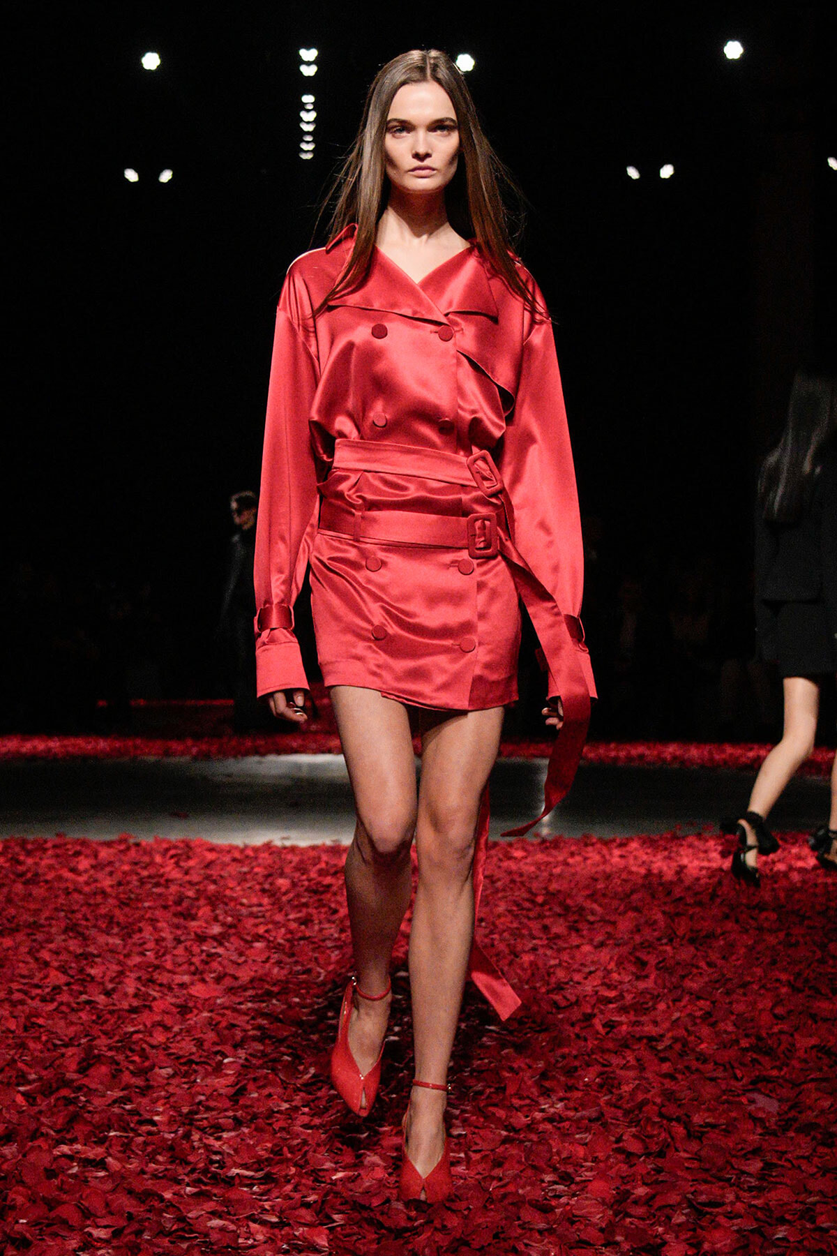 Model walking the runway in a red satin trench dress with belt on a petal-strewn floor.