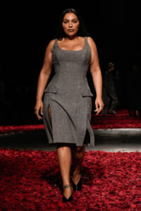 Plus-size model walks the runway in a silver-gray textured dress with side slits, black heels, and red petals on the floor.