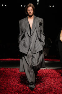 Model walking a runway in an oversized gray pinstripe suit—a double-breasted blazer with wide-leg pants, on a floor covered in red petals.