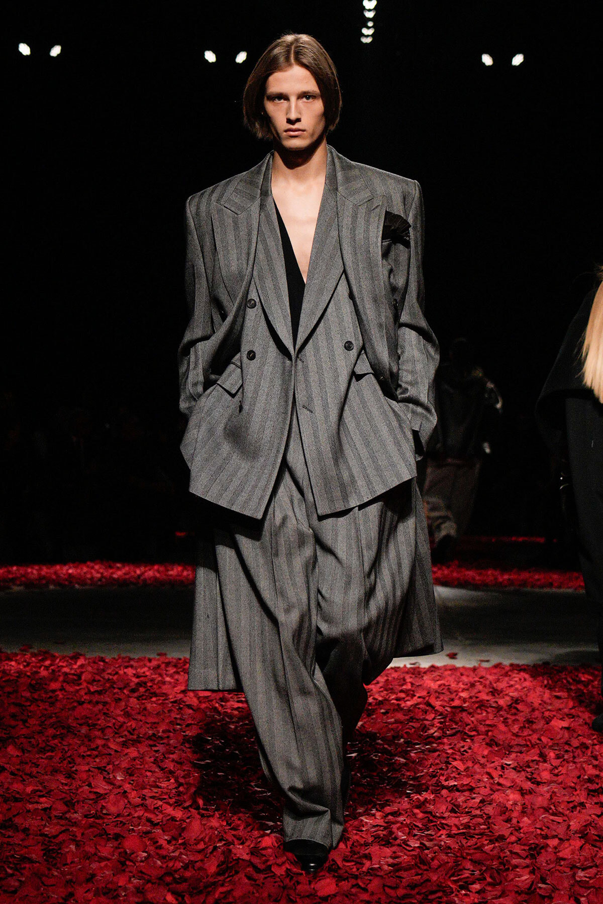 Model walking a runway in an oversized gray pinstripe suit—a double-breasted blazer with wide-leg pants, on a floor covered in red petals.