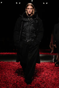 Model walking a runway in an all-black avant-garde outfit with a padded jacket, against a dark backdrop and red petals on the floor.