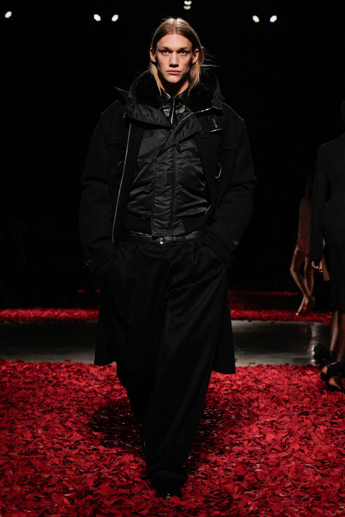 Model walking a runway in an all-black avant-garde outfit with a padded jacket, against a dark backdrop and red petals on the floor.