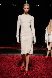 Model walks the runway in a white knit sheath dress with long sleeves, standing on a floor strewn with red petals.