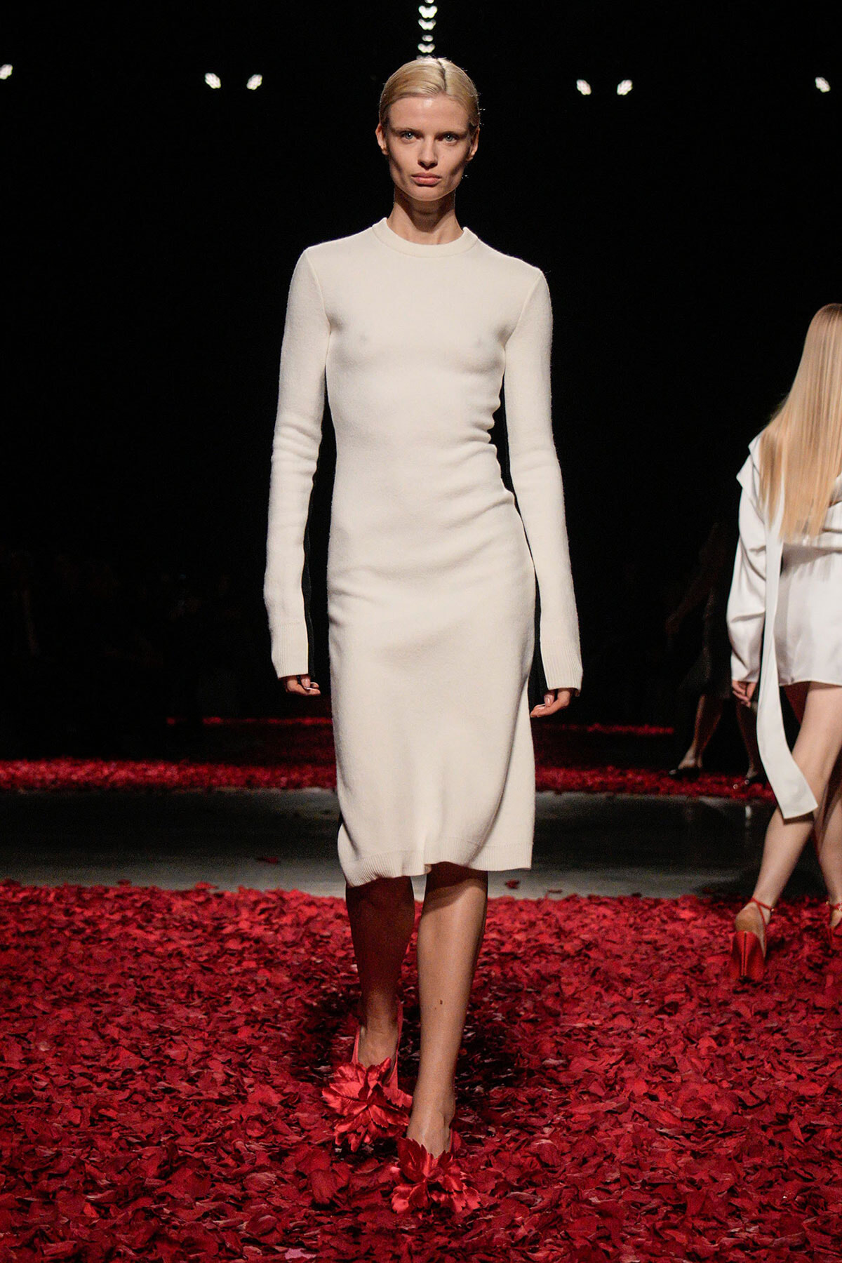 Model walks the runway in a white knit sheath dress with long sleeves, standing on a floor strewn with red petals.