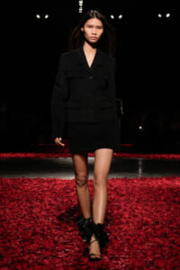 Model walking a runway in a black blazer and mini skirt amid red petals on the floor.