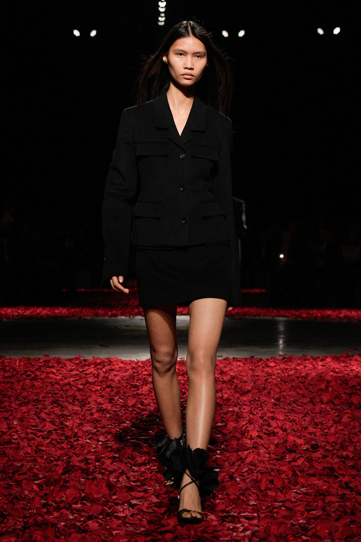 Model walking a runway in a black blazer and mini skirt amid red petals on the floor.