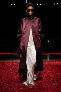 Model walks a runway in a long burgundy satin coat over white wide-leg pants, wearing dark sunglasses, surrounded by red petals on the floor.