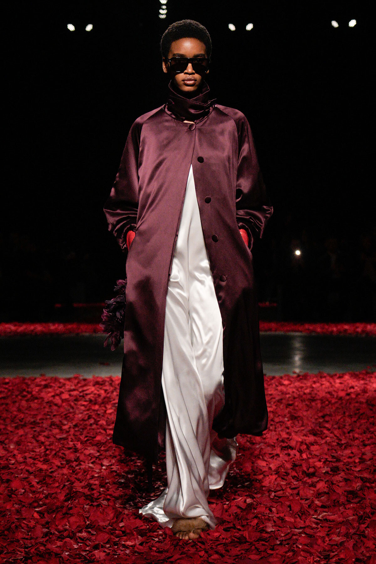 Model walks a runway in a long burgundy satin coat over white wide-leg pants, wearing dark sunglasses, surrounded by red petals on the floor.