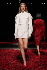 Model walks a fashion runway wearing a white high-neck mini dress and red heels; floor covered in red petals, dark venue in background with another model in a red feathered coat visible behind."