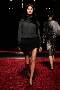Model walking on a runway in a gray and black layered outfit, black high heels, red petal-covered floor. Behind her, another model in sheer graphic dress walks away.