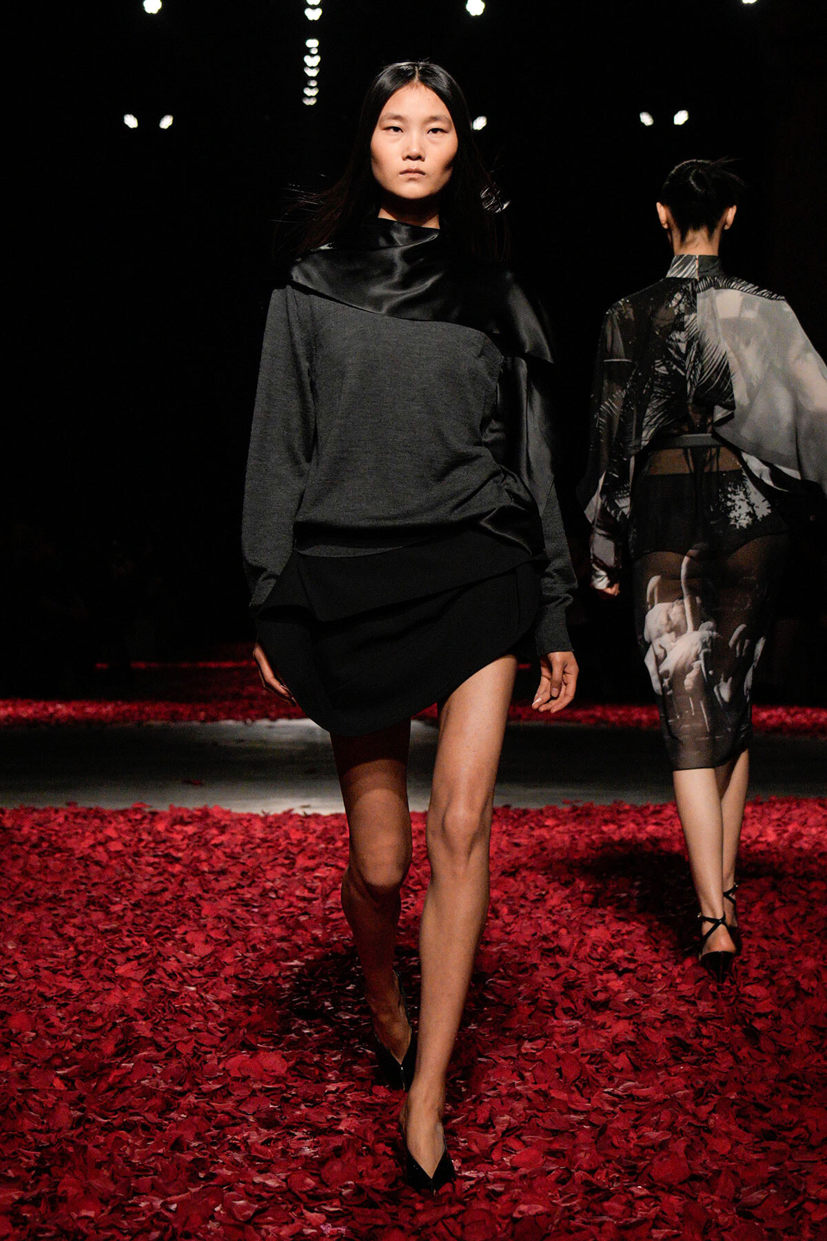 Model walking on a runway in a gray and black layered outfit, black high heels, red petal-covered floor. Behind her, another model in sheer graphic dress walks away.