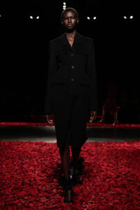 Model walks the runway in a black, knee-length tailored coat with front buttons, on a red petal-covered floor under spotlights; dark background.