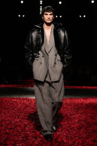 Male model walks a runway wearing a gray herringbone suit with a plunging neckline and a black oversized leather jacket, standing amid red rose petals on the floor.