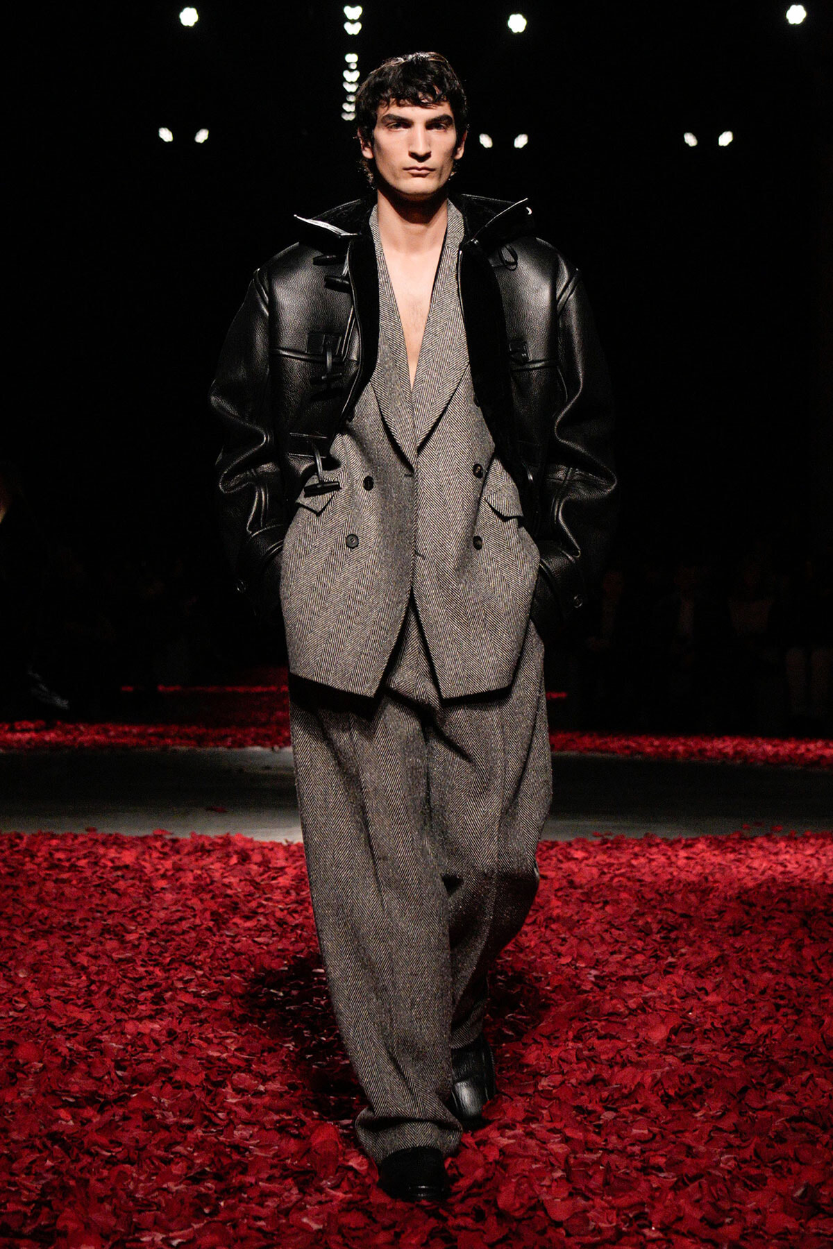 Male model walks a runway wearing a gray herringbone suit with a plunging neckline and a black oversized leather jacket, standing amid red rose petals on the floor.