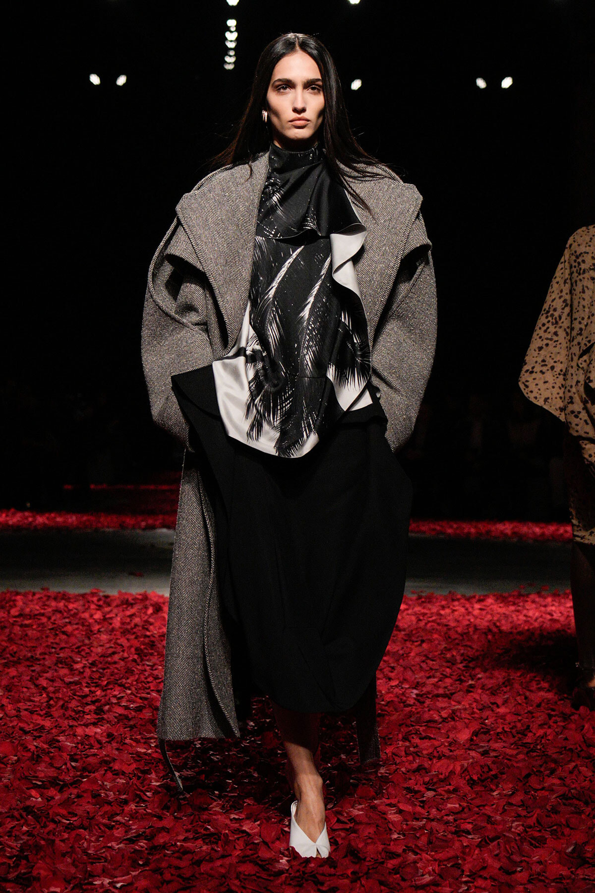 Model walking a runway in a textured gray overcoat over a black-and-white blouse with abstract pattern, wearing a black skirt and white heels on a red petal floor.