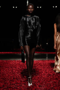 Black-satin long-sleeve mini dress on a model walking a runway surrounded by red petals. White heels complete the look under spotlight.