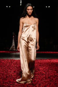 Female model walks the runway in a champagne satin slip jumpsuit on a red rose-petal runway at a fashion show.