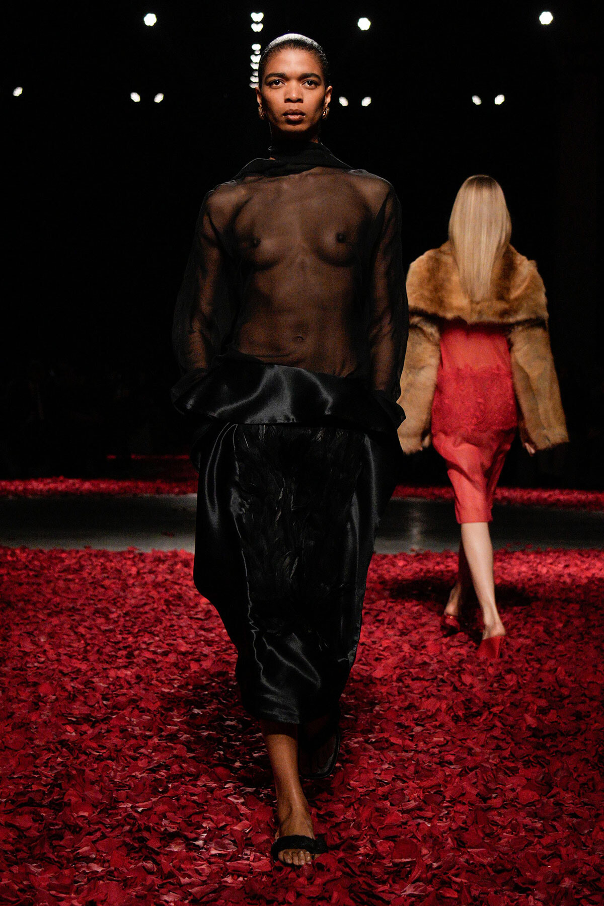 Model walks toward the camera on a runway covered in red petals, wearing a sheer black top and black satin pants.