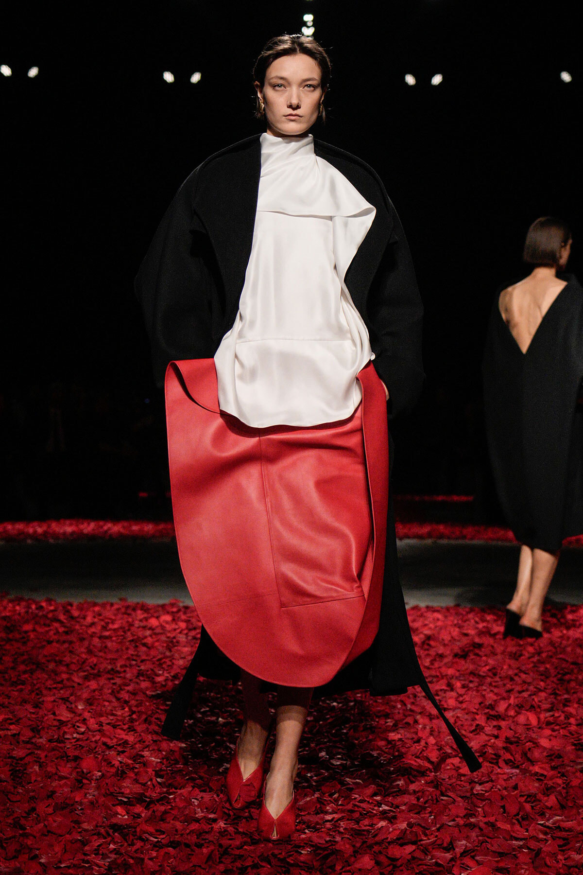 Model walks a fashion runway wearing a black overcoat over a white draped top, carrying a large red tote-like accessory and matching red shoes amid a floor covered in red petals.
